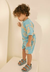 Himmelblaues kurzärmeliges Hemd und passende Shorts mit orangefarbenen abstrakten Mustern; graue Sandalen mit zwei verstellbaren Riemen.