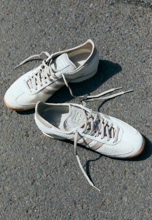 Pár béžových tenisek Adidas s rozvázanými tkaničkami položených na šedém asfaltu na slunci.