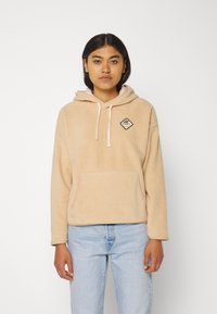 Beige fleece hoodie with drawstring hood, front pocket, and a diamond-shaped logo patch on the left chest. Relaxed fit, long sleeves.