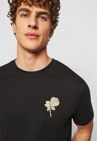 Black cotton t-shirt featuring a beige rose graphic on the left chest area, round neckline, and short sleeves.