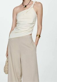 Žena v krémovém topu na jedno rameno s uzlovým detailem, béžových širokých kalhotách, zlatém náramku, náhrdelníku se čtvercovým přívěskem a držící tkanou tašku.