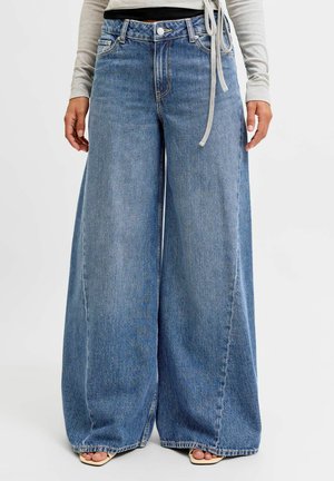 Persona che indossa jeans denim a gamba larga azzurri chiari abbinati a sandali beige con punta aperta e una maglia a maniche lunghe grigio chiaro.