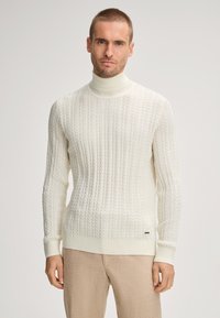 Crème kleurige coltrui met een textuur van kabelbreipatroon, voorzien van lange mouwen en geribbelde manchetten. Gemaakt met lichte bruine broek.