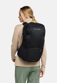 Sac à dos Vaude noir avec une surface texturée, une forme épurée, des bretelles ajustables et plusieurs compartiments zippés. Modèle portant un haut beige clair.