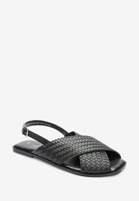 Sandalia negra de tejido con diseño de tiras entrecruzadas, suela plana, tira ajustable en el tobillo y textura de cuero suave.