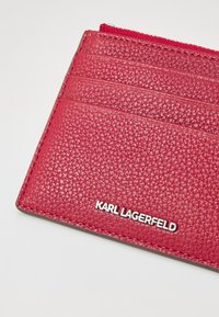 Carteira de cartão em couro texturizado vermelho com múltiplos compartimentos, apresentando um logótipo metálico "KARL LAGERFELD" e um fecho com zíper na parte superior.