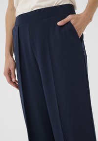 Pantalon large bleu marine avec une texture lisse, doté de poches latérales et de détails plissés à la taille.