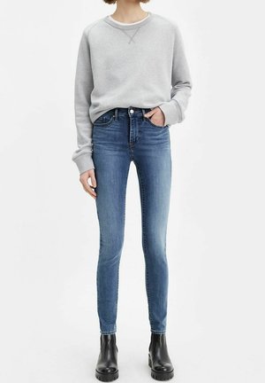 Person iført en lysegrå sweatshirt, blå skinny jeans og sorte ankelstøvler, stående med den ene hånd i lommen mod en hvid baggrund.