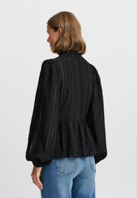 Musthave musta tekstuuriga blousee kõrge kaelusega ja põlvili varrukatega, millel on vertikaalsed triibud ning peplumi alumine äär, koos helesiniste teksadega.
