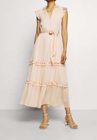 Vestido largo de color rosa claro con mangas y dobladillo acampanados, cierre de botones en el frente, cintura ceñida con cinturón y diseño de capas con textura suave. Estilizado con sandalias de tiras.