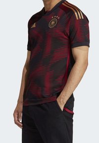 Saksan maajoukkueen jalkapallopaita, musta punaisella geometrisella kuviolla, jossa on kultaiset raidat hartioilla ja vaakuna neljän tähden kera.