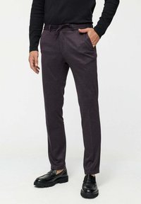 Man wearing dark purple slim-fit trousers with drawstring, black long-sleeve shirt, and black leather loafers, standing with one hand in pocket.