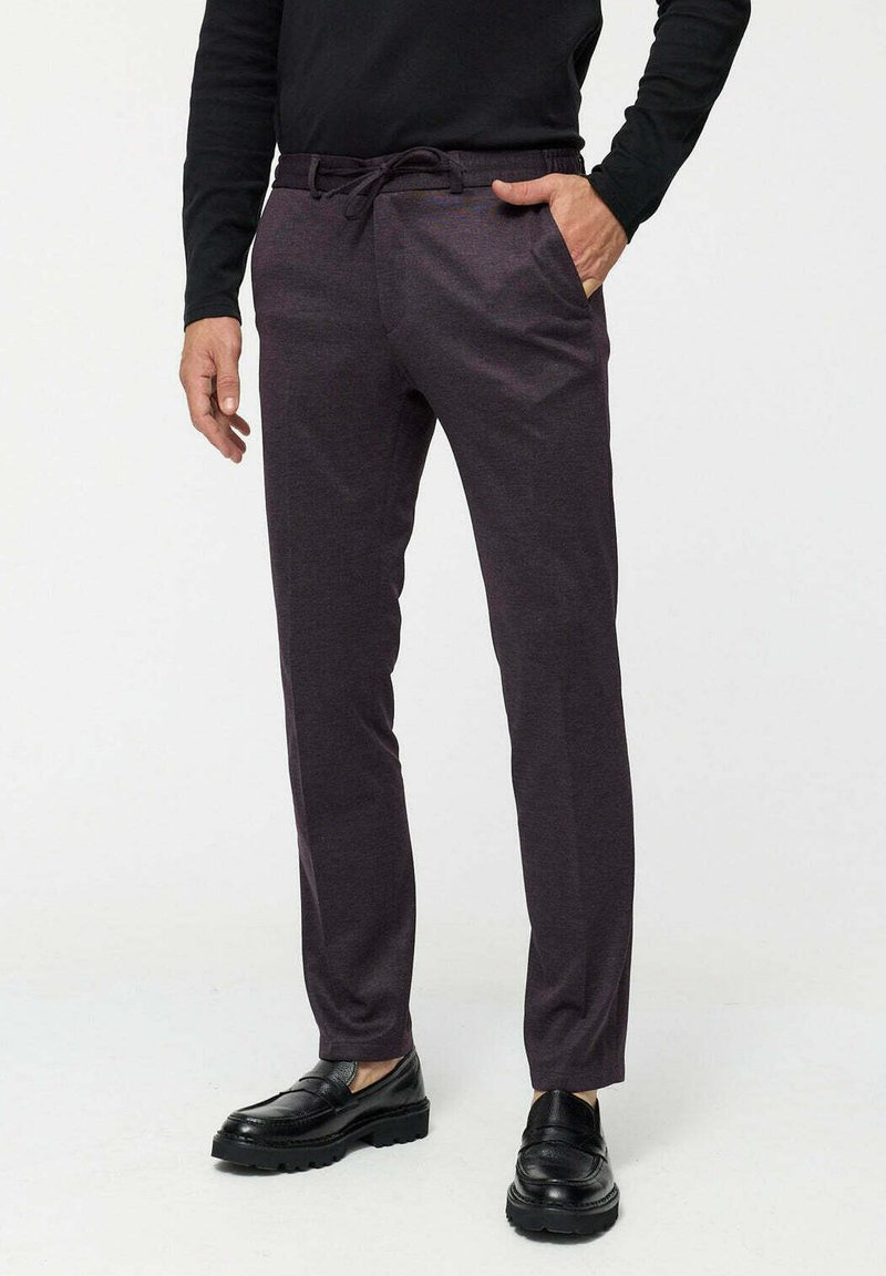 Man wearing dark purple slim-fit trousers with drawstring, black long-sleeve shirt, and black leather loafers, standing with one hand in pocket.