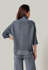 Frau von hinten trägt einen gerippten grauen Rollkragenpullover mit 3/4-Ärmeln und passende weite Hosen vor einfarbigem Hintergrund.