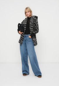 Manteau texturé noir et gris rayé sur un haut noir, associé à un jean bleu à taille haute et large, avec des chaussures noires, tenant un petit sac noir.