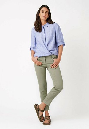 Woman in light blue buttoned shirt with rolled sleeves, olive green pants, and brown sandals standing against white background.