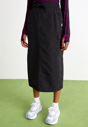 Jupe midi noire avec taille à cordon et poche latérale portée avec un haut à manches longues violet à motif et des baskets blanches sur un sol vert.