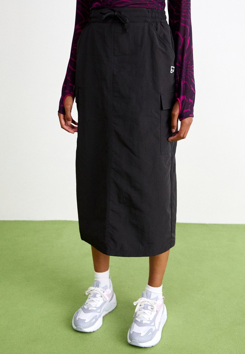 Jupe midi noire avec taille à cordon et poche latérale portée avec un haut à manches longues violet à motif et des baskets blanches sur un sol vert.