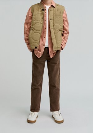 Kind draagt beige gewatteerd vest over rood geruit overhemd, bruine broek en witte sneakers tegen een effen lichte achtergrond.