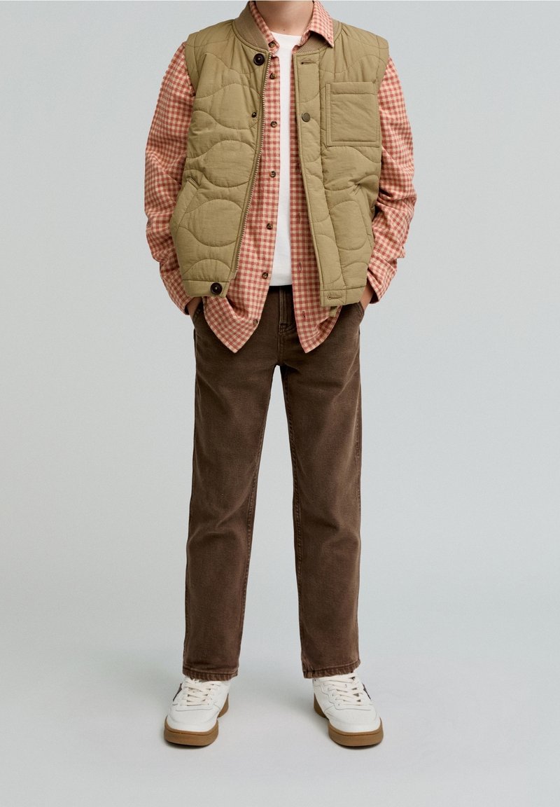 Enfant portant un gilet matelassé beige sur une chemise à carreaux rouge, un pantalon marron et des baskets blanches sur un fond clair uni.