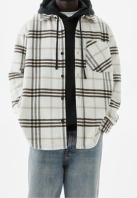 Camicia in flanella con motivo a quadri bianco, marrone e beige; presenta una tasca, chiusura con bottoni ed è sovrapposta a un hoodie nero.