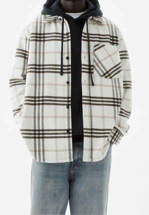 Flanellen overhemd met een geruit patroon in wit, bruin en beige; heeft een zak, knoopsluiting en is gelaagd over een zwarte hoodie.