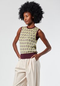 Top senza maniche in maglia verde con un motivo geometrico e scollo e orlo a coste viola scuro, abbinato a pantaloni plissettati color crema.
