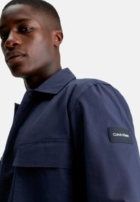Camicia a maniche corte blu navy realizzata in tessuto morbido, dotata di taschino e di un'etichetta nera Calvin Klein sull'avambraccio.