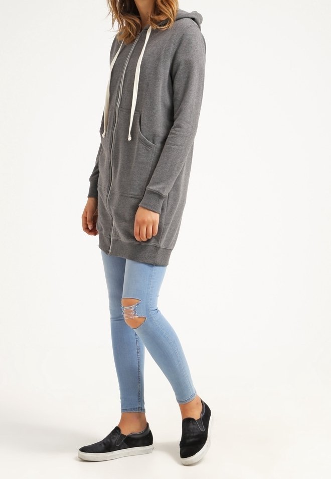 Femme portant un long sweat à capuche gris zippé, un jean skinny bleu clair déchiré, et des baskets noires sans lacets, debout sur un fond blanc.