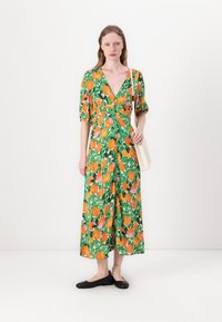 Grünes Blumenkleid mit großen orangefarbenen und pinken Blumen, gepufften Ärmeln, einem tiefen V-Ausschnitt und Knöpfen vorne. Kombiniert mit schwarzen Ballerinas.