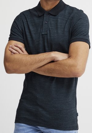 Homme portant un polo à manches courtes gris foncé et un jean bleu clair, les bras croisés sur la poitrine.