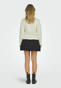 Bijela pletena sweatshirt s teksturiranim uzorcima, u kombinaciji s crnom plisiranom suknjom i crnim čizmama do gležnja. Vidljiv stražnji pogled i frizura.