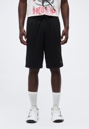 Homme portant un short de sport noir, des chaussettes blanches, des baskets blanches et un t-shirt blanc à motif avec le texte "HOOPS" et un dessin de basket.