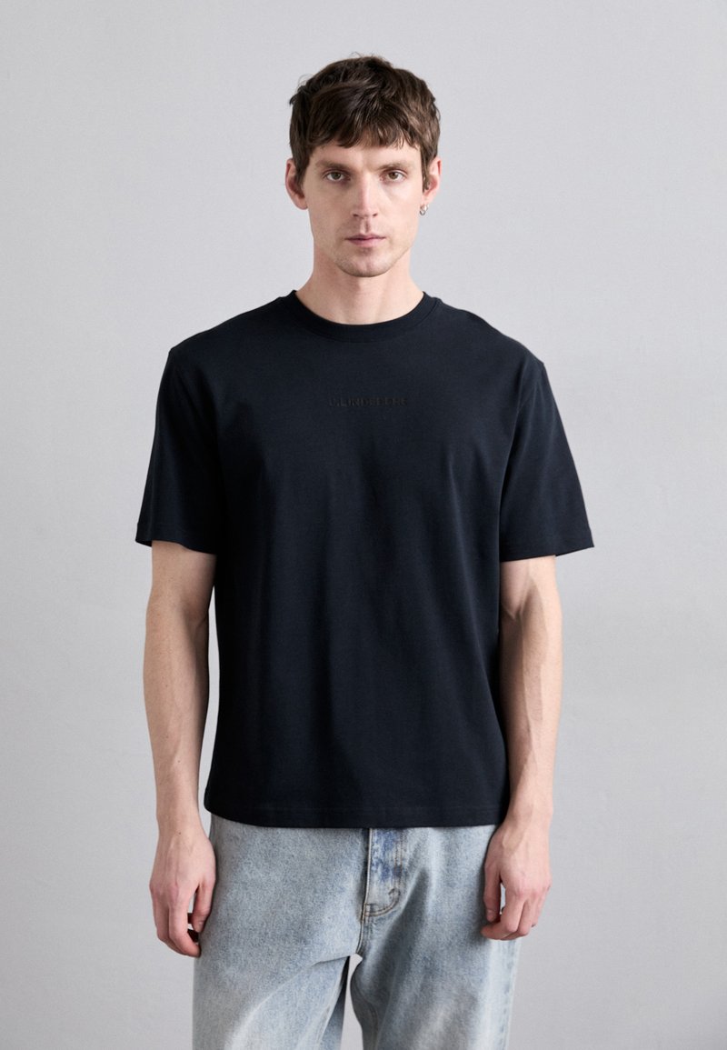 Camiseta de algodón negra de manga corta, con cuello redondo y logo en relieve en el pecho. Combinada con unos jeans de color azul claro. Diseño simple.
