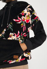 Czarna bluza z kapturem z kwiecistym wzorem w różowym, białym i zielonym kolorze. Posiada kieszeń kangurkę oraz naszywkę marki Roxy. Aksamitna tekstura z przodu.