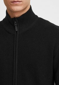 Sudadera negra con cremallera, cuello alto acanalado, tela de punto texturizada y detalle de cremallera frontal. Adecuada para prendas en capas.