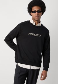 Sudadera negra con "MORATO" impreso en gris reflectante, con un cuello redondo y puños acanalados. Lleva puesto encima de una camisa blanca con cuello.