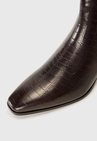 Bottine marron en cuir embossé de crocodile avec un bout pointu et un design épuré. Talon haut et texture lisse, avec un rebord renforcé.