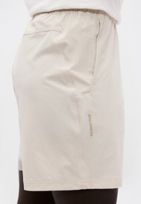 Μπεζ σορτς με ελαστική μέση και πλαϊνή τσέπη, με το λογότυπο "SALOMON" στο δεξί μηρό, φορεμένα πάνω σε λευκό φόντο.