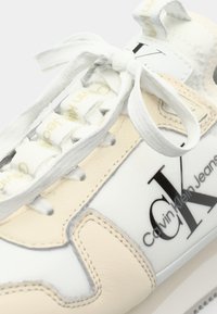 Adidașii Calvin Klein din piele albă și bej, cu un finisaj neted, sunt caracterizați de un logo și șireturi plate albe. Talpa din cauciuc cu model este vizibilă.