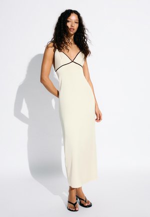 Femeie cu păr lung și creț, purtând o rochie maxi crem fără mâneci, cu margini negre și sandale negre, stând în picioare pe un fundal alb.