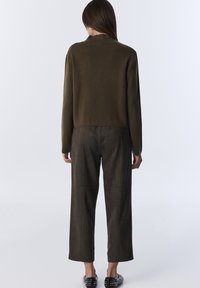 Kobieta stojąca tyłem, ubrana w ciemnooliwkowy sweter, ciemne szerokie spodnie i czarne slip-on'y z ćwiekami na jednolitym tle.