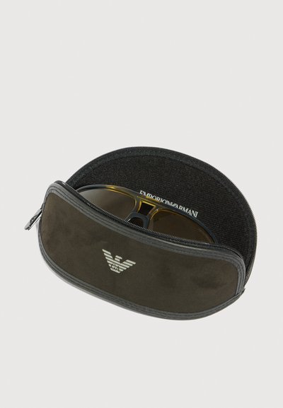Melnā, ar rāvējslēdzēju aizverama saulesbrīļu soma ar Emporio Armani logotipu, nedaudz atvērta, atklājot ūdenspārredzamas brūnas saulesbrilles iekšā, uz balta fona.
