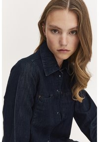 Cămașă din denim de un albastru închis, cu guler clasic, închidere cu nasturi și buzunare frontale. Dispune de detalii de cusătură subtile și un material ușor texturat.
