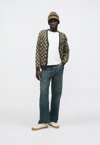 Checker-patterned cardigan in brown and beige, white t-shirt, flared denim jeans, and a multicolored knit beanie. White sneakers with gum soles.