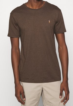 Homme portant un t-shirt marron à manches courtes et col rond avec un petit logo brodé sur la poitrine et un pantalon beige.