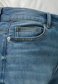Φωτεινό μπλε τζιν με ορατές ραφές μπροστινής τσέπης και μικρή τσέπη για κέρματα, φορεμένα με λευκό πουκάμισο που είναι μέσα από το παντελόνι.