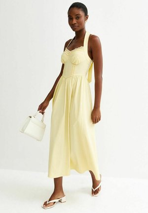 Kobieta w jasnopomarańczowej sukience midi na szyję, trzymająca białą torebkę i nosząca białe sandały na obcasie, stojąca na białym tle.