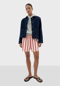 Veste en jean, bleu foncé, avec deux poches avant, associée à un short rayé rouge et blanc. Des tongs noires complètent la tenue.