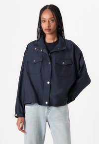 Giacca cropped blu realizzata in tessuto leggero, con due tasche anteriori con pattina, bottoni a pressione e colletto rialzato con una leggera lucentezza.